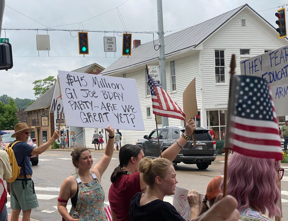 Woman holding up a sign reading “$45 Million GI Joe BDay Party…Are We Great Yet?”