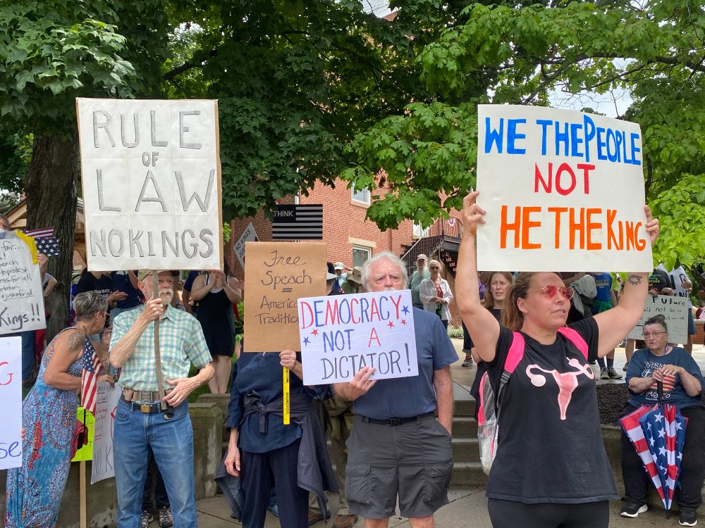Three protesters holding signs: “Rule of Law,” Democracy Not A Dictator,” and “We The People Not He The King”