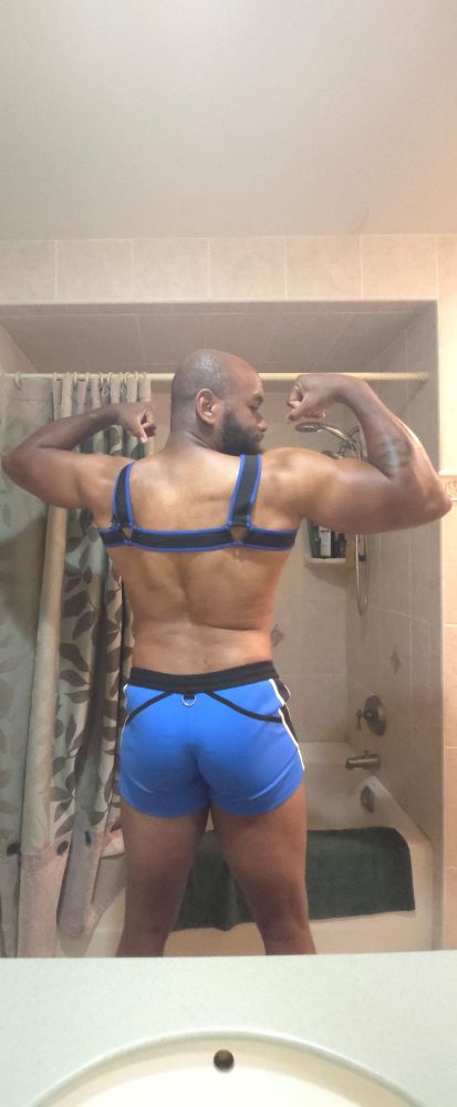 Flexing both biceps with back turned to camera. Wearing blue shorts and similar colored harness