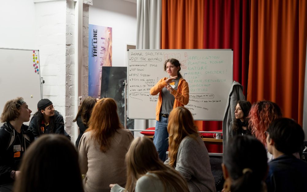 A woman pitching a game idea with a whiteboard behind her in front of a group of people.