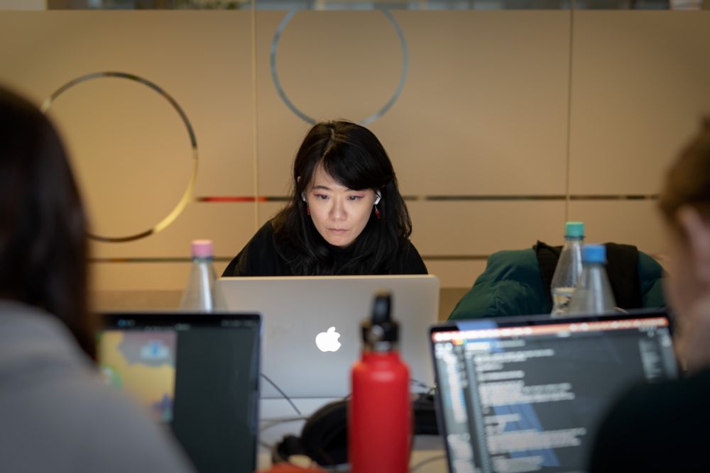 A woman sitting at a table, looking at a Macbook screen in concentration while being surrounded by other hardware and waterbottles.