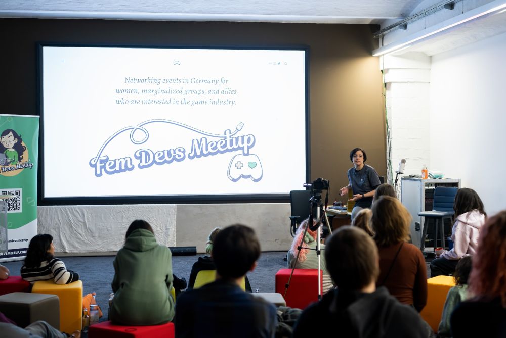 A woman holding a presentation about the FemDevsMeetup initiative on a projector screen in front of a group of people.