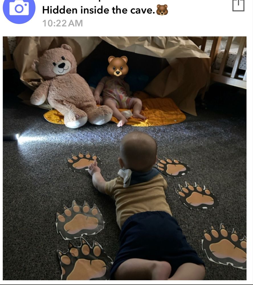 a baby lays on his stomach between two rows of paper bear paw prints. In front of him is a stuffed bear and another baby sitting in a cave made of brown paper 