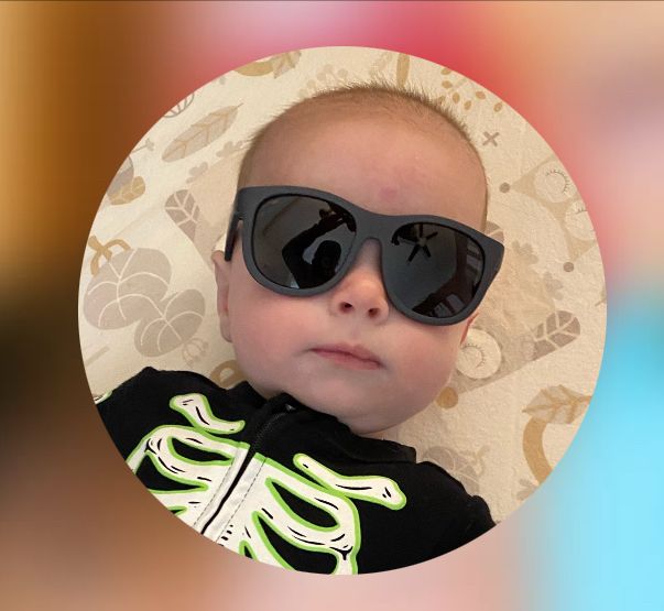 a round-faced baby with fuzzy brown hair is wearing a skeleton baby onesie with sunglasses. he has a serious look on his face