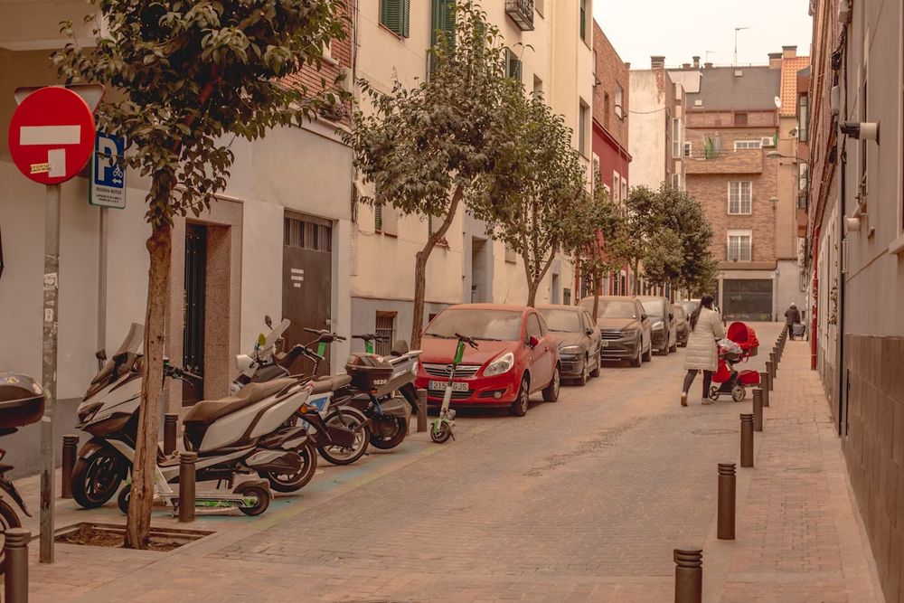 Calle residencial de Madrid. Cerca de un tercio del espacio se dedica a estacionamiento de vehículos a motor. Más o menos la mitad del espacio se dedica a que puedan rodar los mismos. El espacio peatonal está perimetrado con bolardos para los que los vehículos a motor no estacionen y buena parte de la identidad visual de la calle son señales de tráfico.