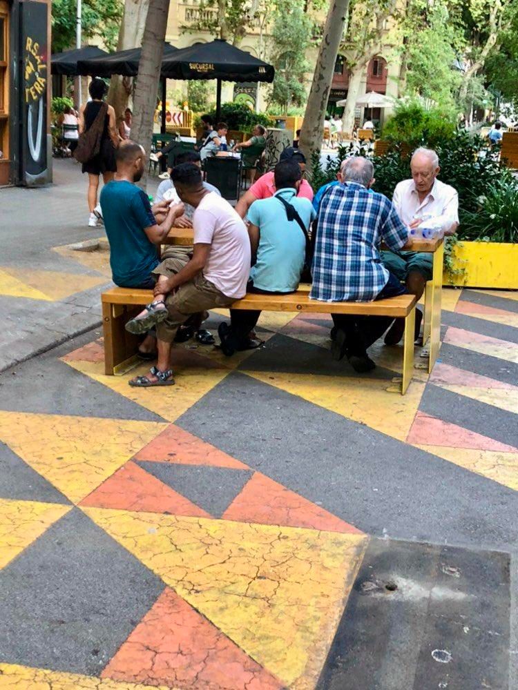 La imagen muestra lo que ocurre cuando se recupera parte de la ciudad ocupada por el coche y se le da a la ciudadanía.

Escena al aire libre con un grupo de personas sentadas en un banco largo de madera y metal. El banco está ubicado en un área pavimentada con un patrón geométrico de triángulos amarillos, naranjas y grises. Alrededor del grupo, se ven árboles y un espacio con mesas y sombrillas de un café o restaurante. En el fondo, hay más personas sentadas en las mesas y algunas caminando. El ambiente parece ser relajado


