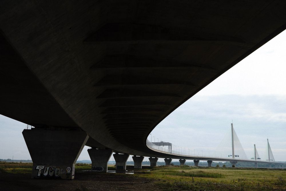 The underside of a great concrete bridge sweeps across the top of the frame. It is painted in shadow, held aloft by austere pillars rising above a flat wasteland.
As the sweep rolls away to the right of frame, contracting to a fine line, tall, sail-like supports reach into a grey sky. It is a vast suspension bridge across the river Mersey, viewed from below.
