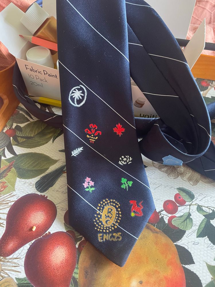 A navy tie with a fine white stripe. Which has now had the Rugby World Cup 2025 logo painted on it, along with the logos of the teams in pool c: NZ, Japan, Ireland and a much less legible Spain and pool b: Fiji, Wales, Scotland and Canada. 
