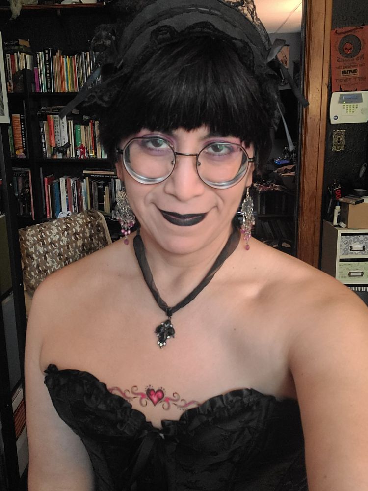 Photograph of a feminine presenting moderately fair skinned person with short dark hair and a black gothic hairpiece trimmed with lace. The person has thick black glasses, and purple eye makeup. They are looking ahead at the camera with a close mouthed smile and black lipstick. There is a black fabric necklace with a black onyx cross their their neck. They are wearing a black sweetheart corset with a pink heart lined temporary tattoo in the center of a flat chest pushed up by the corset. They are in front of a bookshelf and doorway into an office behind them.