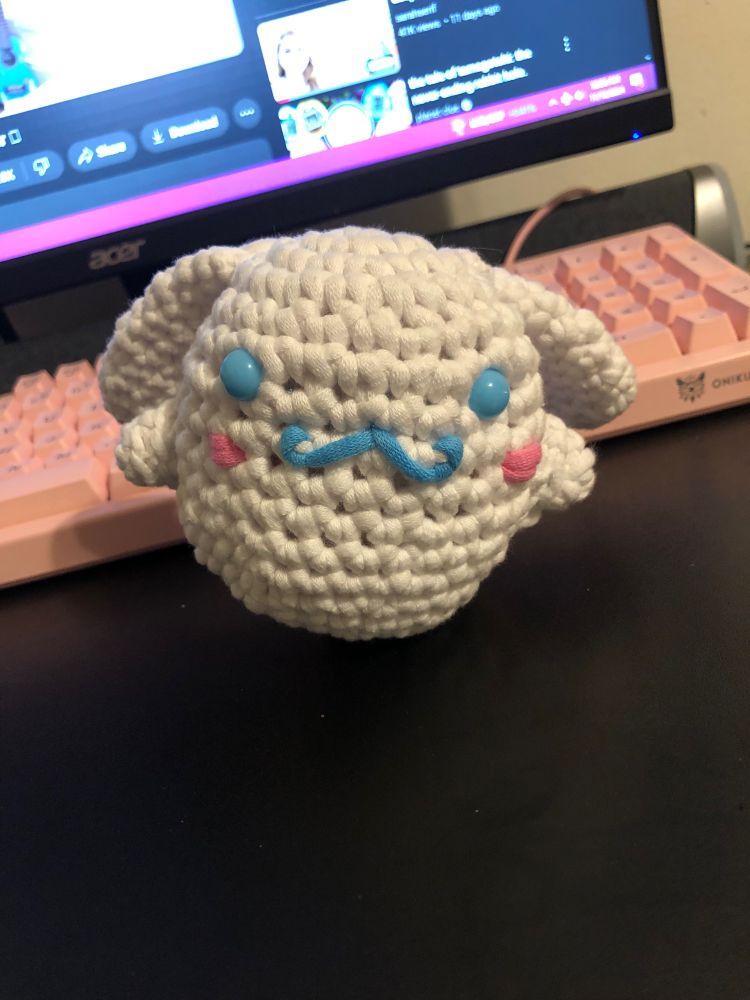 A crocheted cinnamon roll from Sanrio. He’s white in an egg like shape with long ears a blue snap eyes and M curled mouth. With little pink blush lines . Background is a black desk with a pink keyboard 
