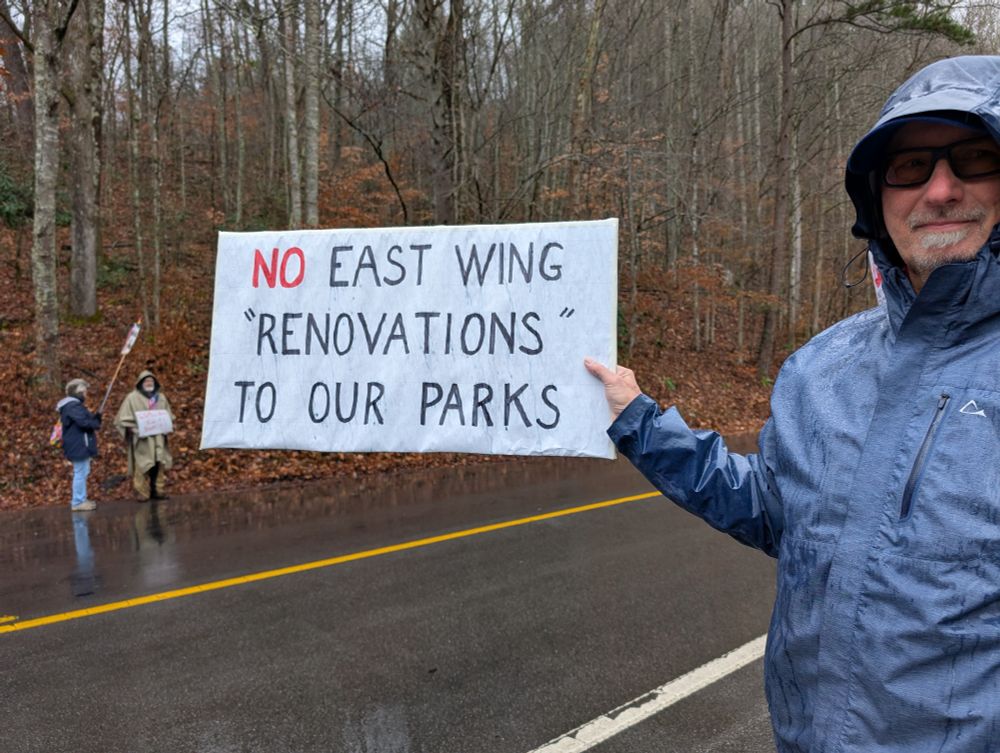 Two protesters visible in the background across the street from one protester who is smiling, wearing a wet rain coat with the hood up, and a pair of glasses. He's holding a white sign with mostly black letters except for the word "no" which is written in red. The sign reads, "no East Wing "renovations" to our parks"