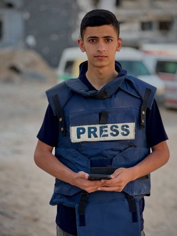 Musab al Sharif wearing the classic blue PRESS vest. He is young and malnourished, a stringy youth with kind, sad, and steadfast eyes.