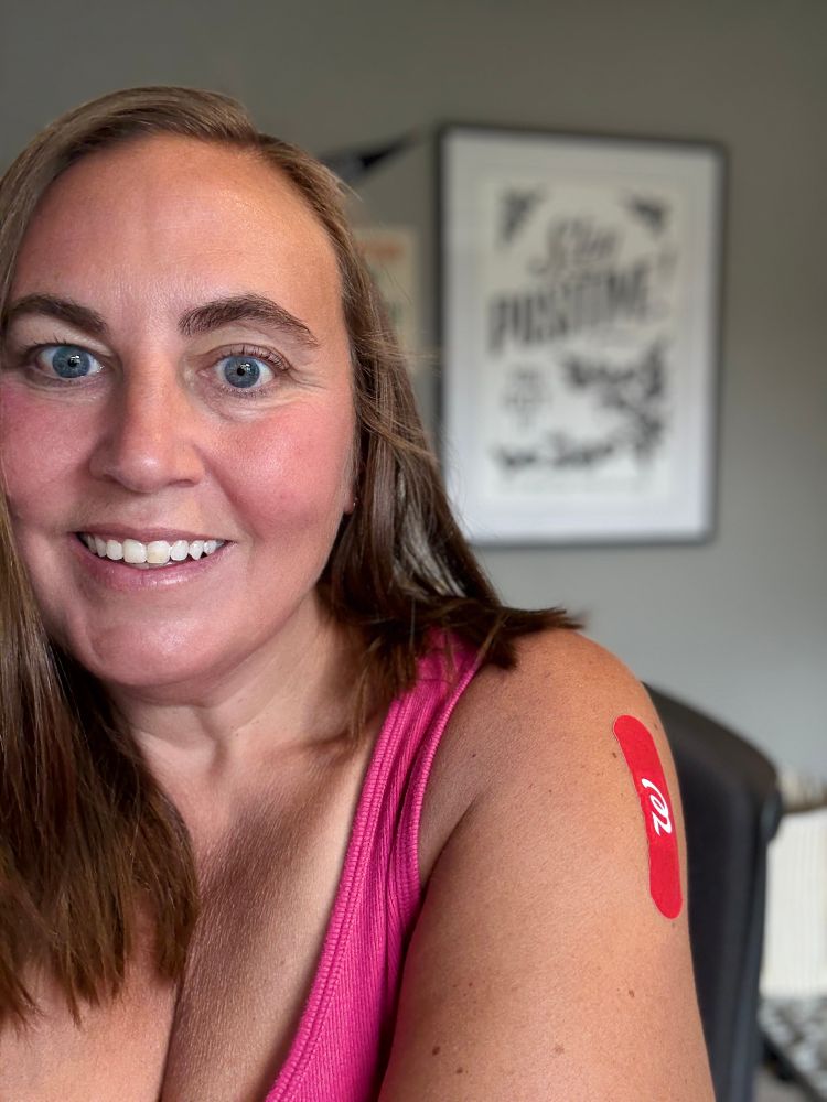 Selfie of woman in a pink tank top with a red Walgreens bandaid on her upper arm. A “Stay Positive: You Got It” Labor Camp Report poster commemorating the day Trump got COVID in October 2020 is in the background. 