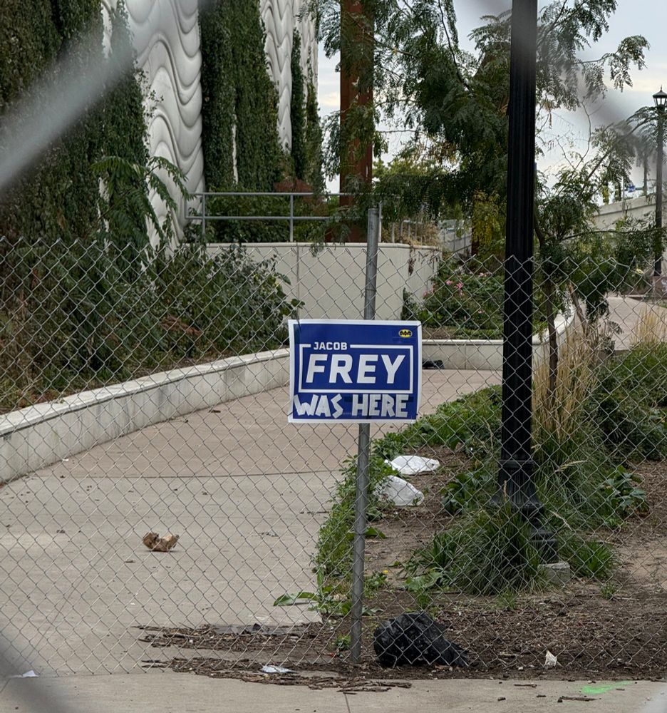 A blue sign with white writing reads Jacob Frey was here through many layers of chain link fence. 
