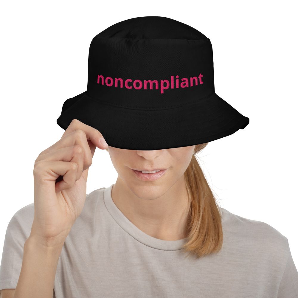 A black bucket hat with a humorous embroidered design on the front. The design features one line of text: "noncompliant" in a large, bold, hot pink font. The hat is shown on a female model, close up. Also comes in blue - promo for Etsy shop.