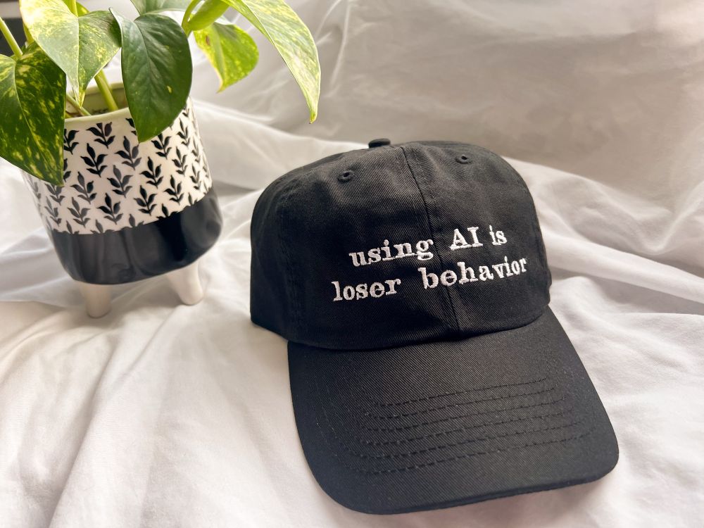 black baseball cap that says “using AI is loser behavior”