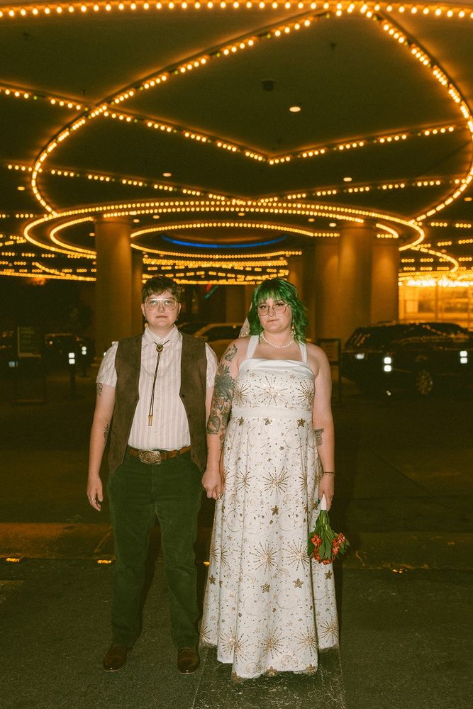 lesbian couple holding hands standing in front of a golden light fixture in vegas. it’s dark outside and the picture was taken with flash. on the left, joey is wearing a brown vest, light shirt, bolo tie, and green pants. on the right, noelle has green hair and is wearing a white/gold celestial wedding dress, and is holding a small red bouquet of flowers