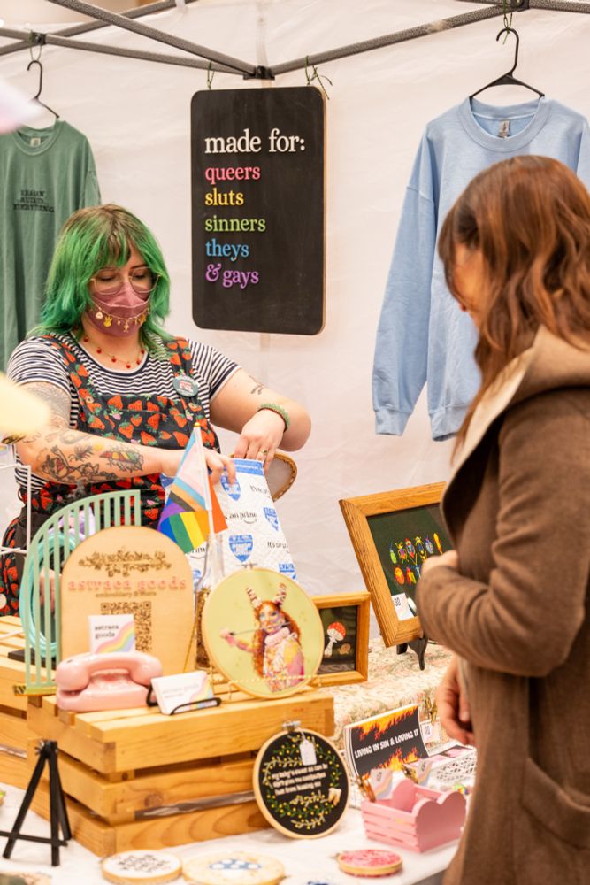 noelle packaging up an embroidery hoop for a customer at an art market. they’re wearing a strawberry jumpsuit, a mask with a gold mask chain, and they have green hair and tattoos. their booth has a rainbow sign that says “made for: queers, sluts, sinners, theys & gays”
