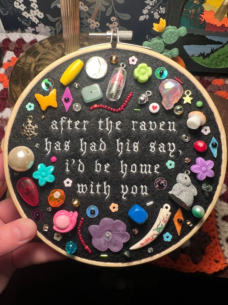 embroidery hoop with chaotic charms/trinkets surrounding a lyric from hozier: “after the raven has had his say, i’d be home with you”