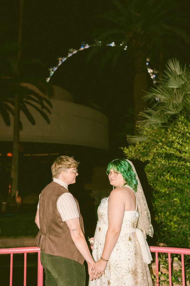 lesbian couple holding hands standing in front of a pink fence with a ferris wheel in the background. it’s dark outside and the picture was taken with flash. on the left, joey is wearing a brown vest, light shirt, and green pants. on the right, noelle has green hair and is wearing a white/gold celestial wedding dress