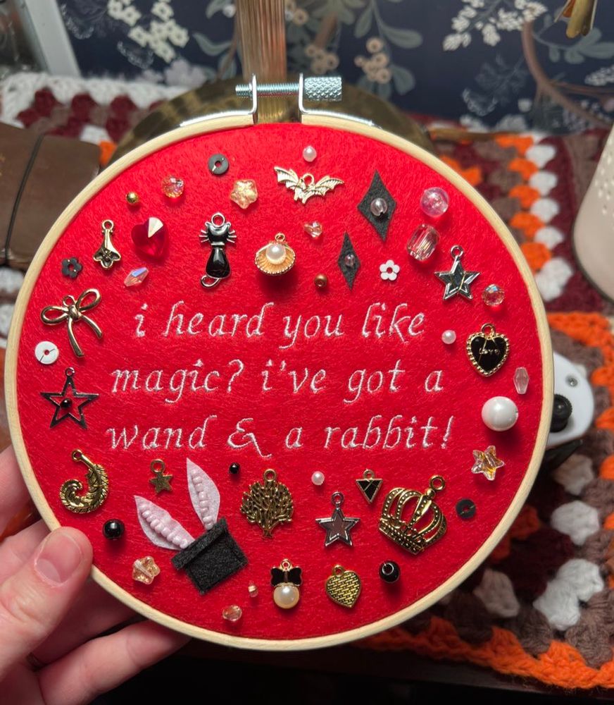embroidery hoop with red fabric that says “I heard you like magic? i’ve got a wand and a rabbit!” in white thread. it’s surrounded by gold, white, and black charms/beads/embroidered details  