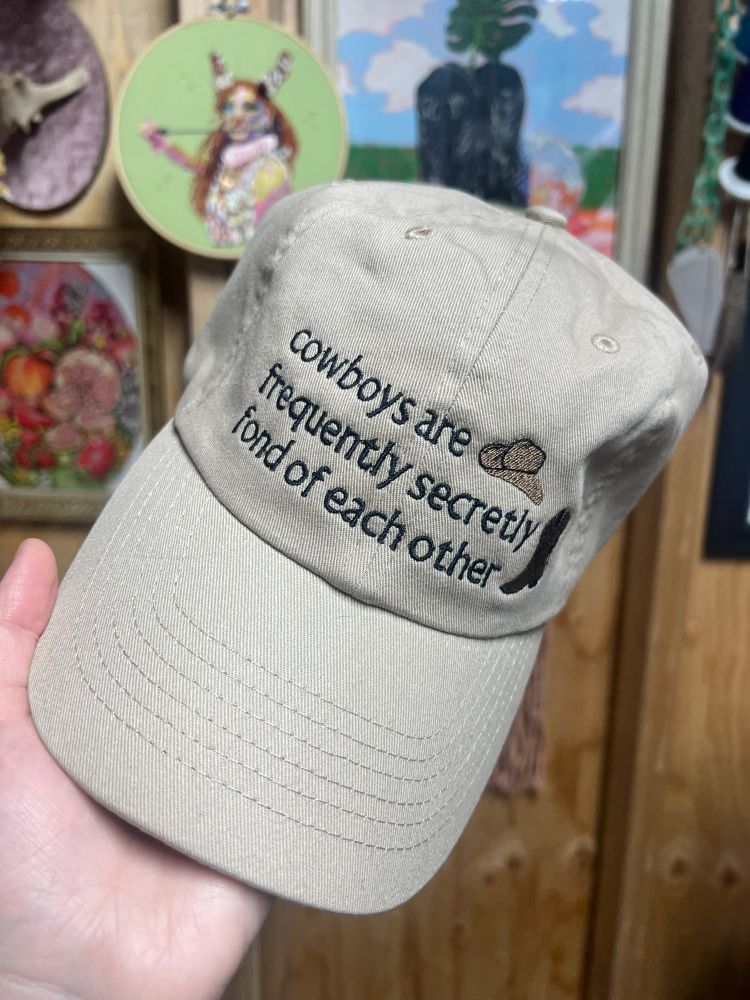 khaki baseball cap with lyrics from a willie nelson and orville peck song: “cowboys are frequently secretly fond of each other”. there’s a cowboy hat and a cowboy boot on the right side of the design