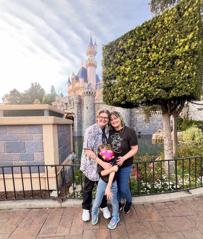 noelle, their wife, and their daughter standing in front of the disneyland castle