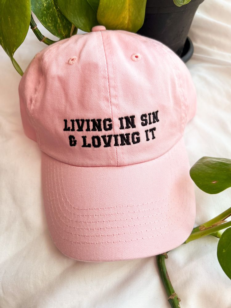 pink baseball cap that says “living in sin and loving it” in black text. there’s a plant head as a prop