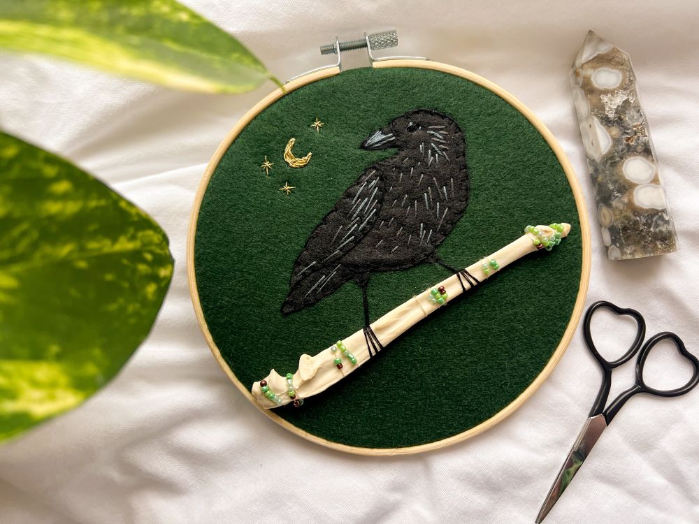 an embroidery hoop with a green felt background. there’s a gold moon and stars on the left side with a black felt crow looking towards the moon. the crow is holding onto a raccoon bone, and the bone has beaded moss along it.