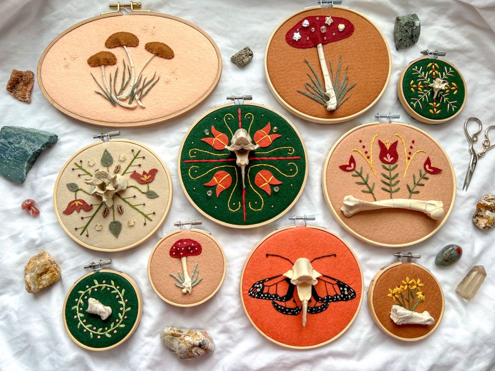 various sizes of embroidery hoops with different colors of felt, bones, and embroidered detail on each. all have earth tones and some are folk-inspired. there are crystals and gold scissors around the edges as props