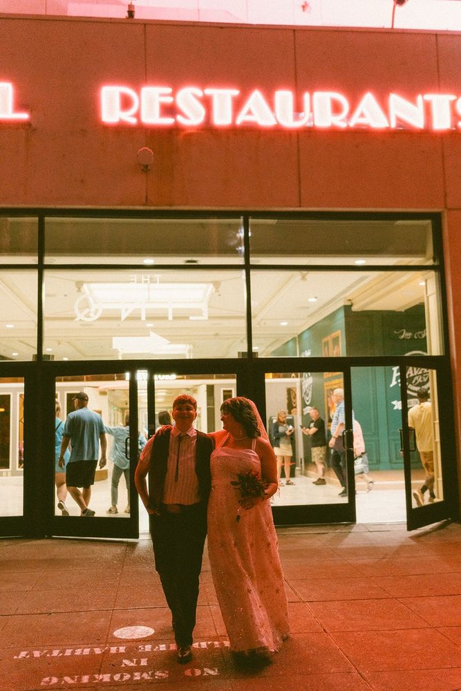 lesbian couple walking towards the camera with pink neon lights behind them. on the left, joey is wearing a brown vest, light shirt, and green pants. on the right, noelle has green hair and is wearing a white/gold celestial wedding dress