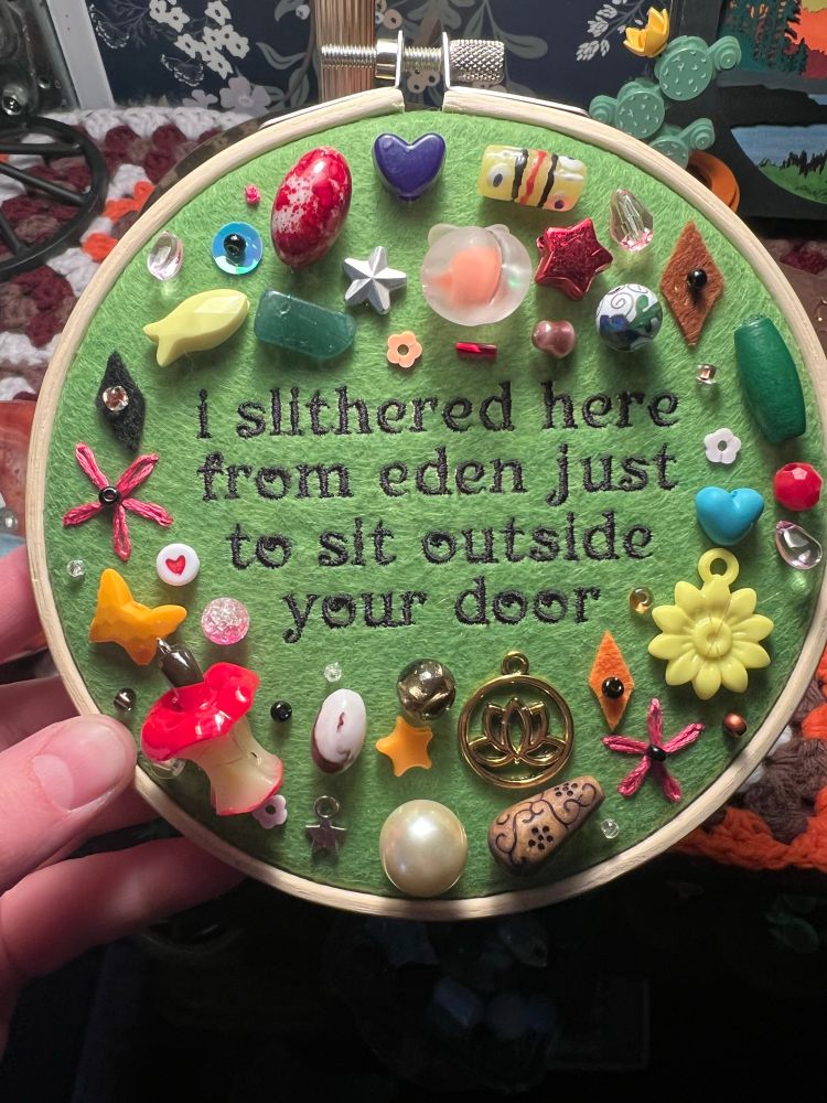 embroidery hoop with chaotic charms/trinkets surrounding a lyric from hozier: “I slithered here from eden just to sit outside your door”