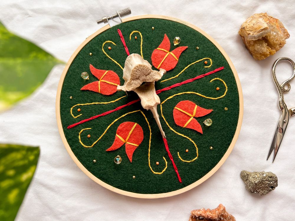 6” wooden embroidery hoop. forest green felt fabric background with orange flowers, yellow and red embroidered details, and beaded accents in a folk-inspired style. features an ethically-sourced coyote vertebrae