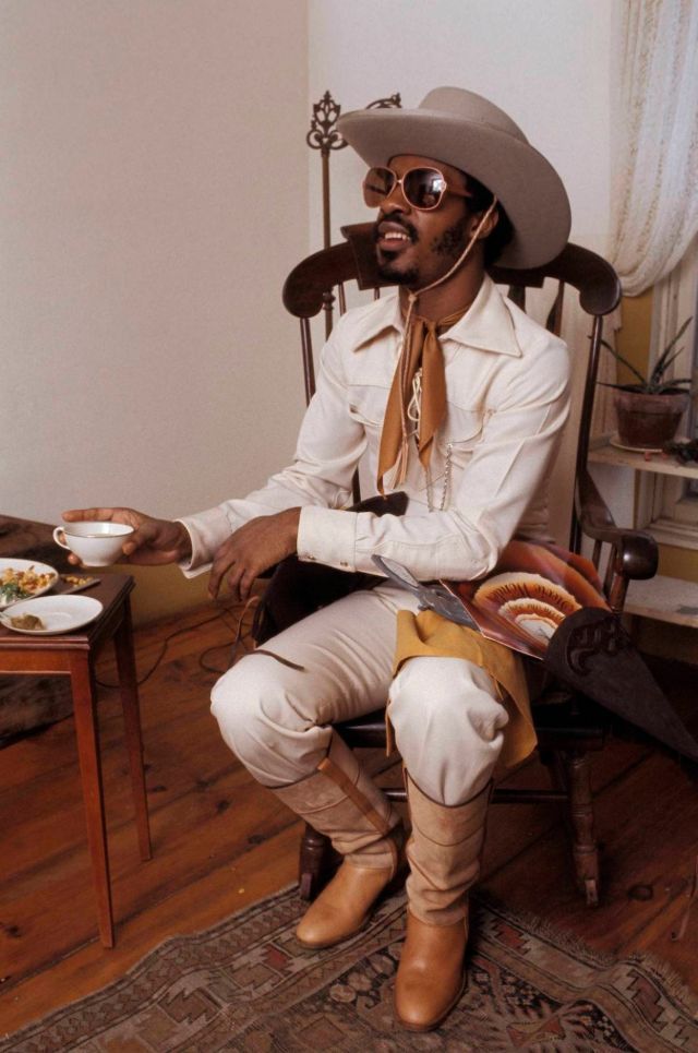 Color photo of Stevie Wonder in a cowboy costume sitting at a small table with a cup of tea in his hand.