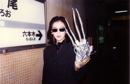 Color photo of Kei Fujiwara standing in a subway station.  She's wearing sunglasses, a dark coat, and a prop robot hand over her real hand.