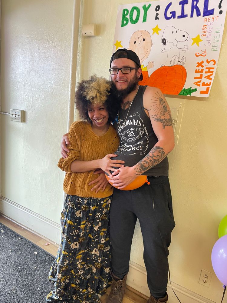 leah and alex, leah is a short black woman with a afro with part of the front dyed blonde. and alex is a tall white man with glasses holding a balloon in his shirt, pretending hes the one thats pregnant.