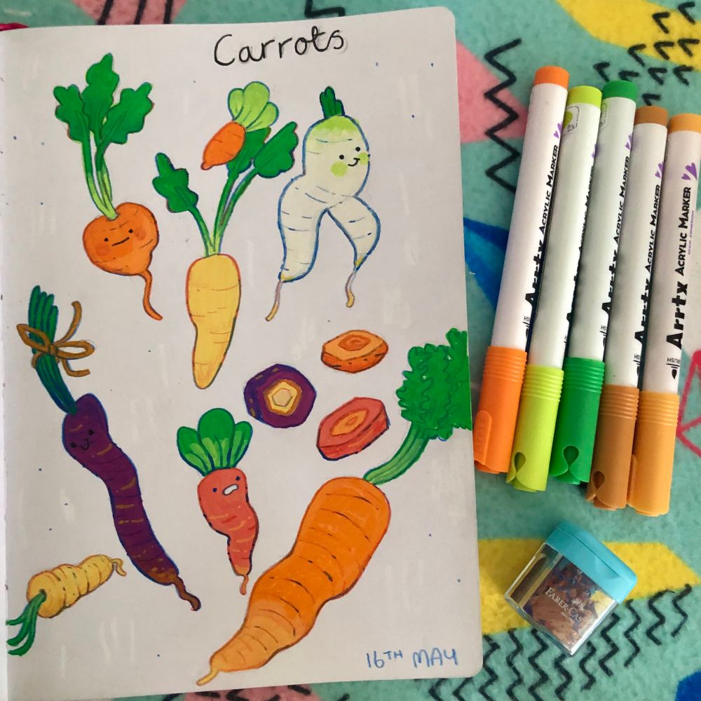 An open sketchbook page next to a handful of paint markers and a pencil sharpener. The page is full of red, orange, yellow, white and purple carrots! Some have little faces drawn on them :o)