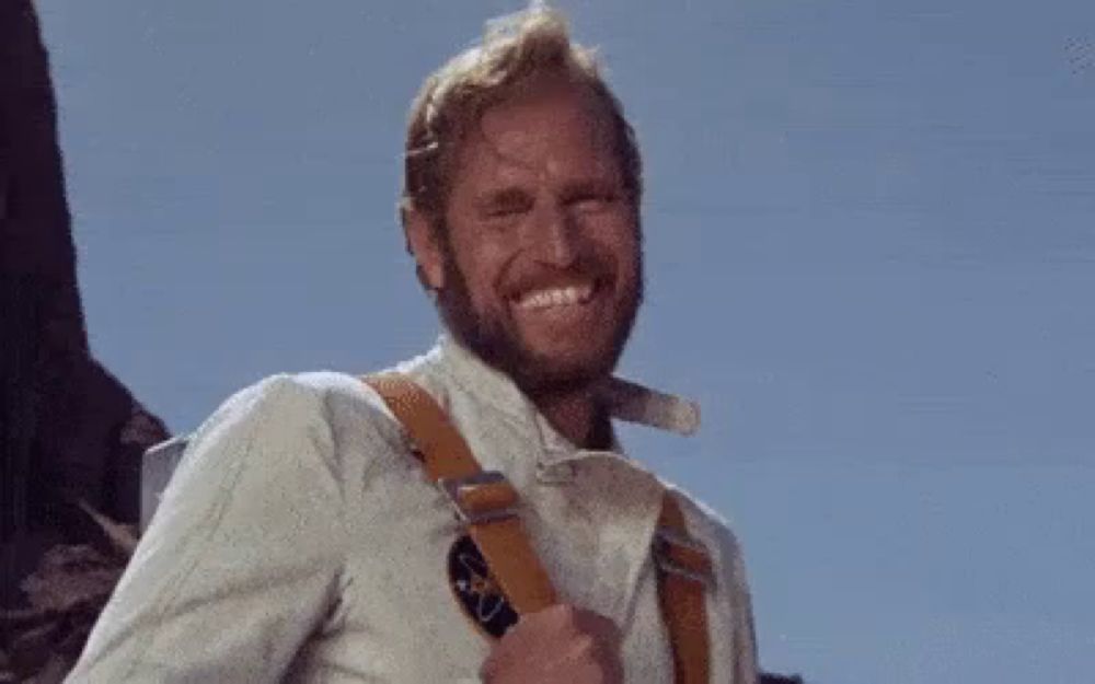 a man with a beard and suspenders is smiling while standing in front of a mountain .