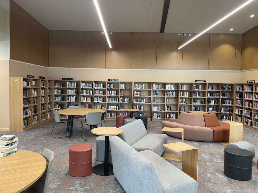 Library space with timber bookshelves around the walls, tables and chairs on the left and soft seating in greys, pink and red in the centre