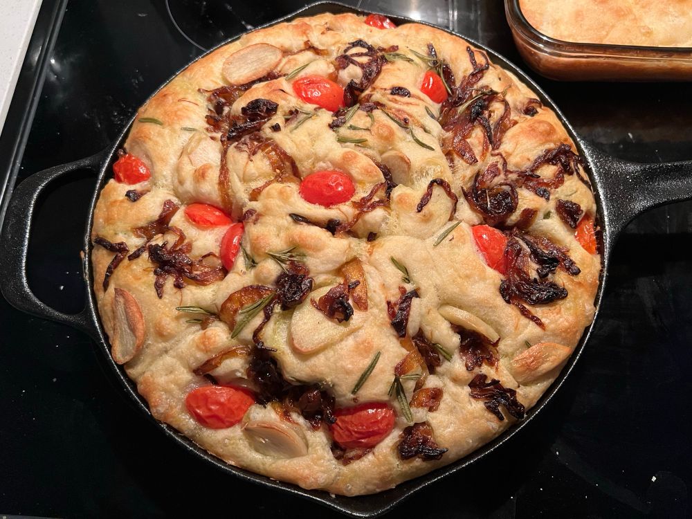 focaccia in a cast iron skillet. There are slices of elephant garlic, cherry tomatoes, onions I caramelized the night before, and fresh rosemary sprinkled on top