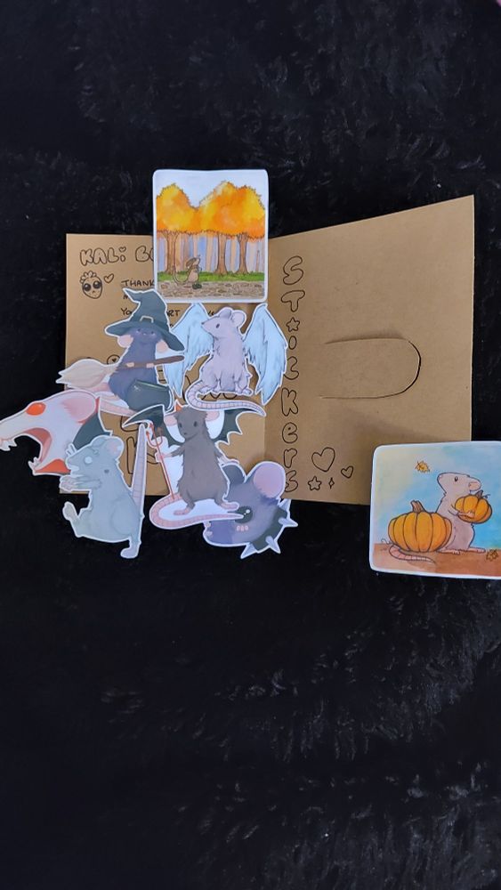 the brown envelope on the black fuzzy background is now opened. an assortment of rat stickers are scattered across the pages. a strawberry with pleading eyes is drawn on the envelope. it says STICKERS vertically on the 2nd page, which also has a cutout sleeve for the stickers to reside in. far right sticker is a rat on a brown surface with a blue background, holding a smaller pumpkin and standing next to a large pumpkin. the rat is looking to the left. top sticker: an autumn forest scene where a rat is walking along the path. there's a blue rat with a witch hat and a broomstick, and light grey rat with white angel wings, a dark grey rat with black devil wings and a red pitchfork, a greyish blue rat face with a spiked collar necklace, a grey zombie rat with its tail pointed straight up and its arms outstretched in front of them as they "march" to the left. an albino vampire rat with white face and red eyes 