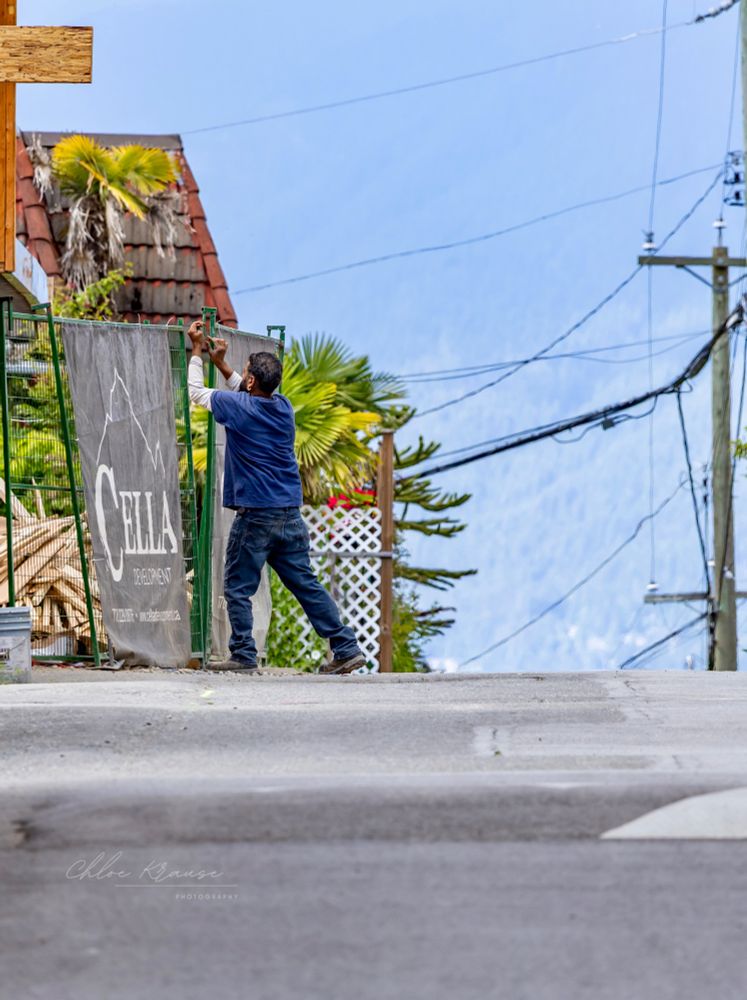 A person wearing denim working to attach a construction fence in front of a house. You can see hazy trees on a mountain in the background. 