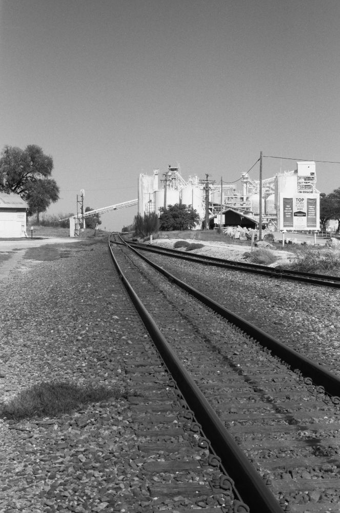 B&W photo of railroad tracks disappearing into the distance, next to a faraway collection of towers and conveyor belts