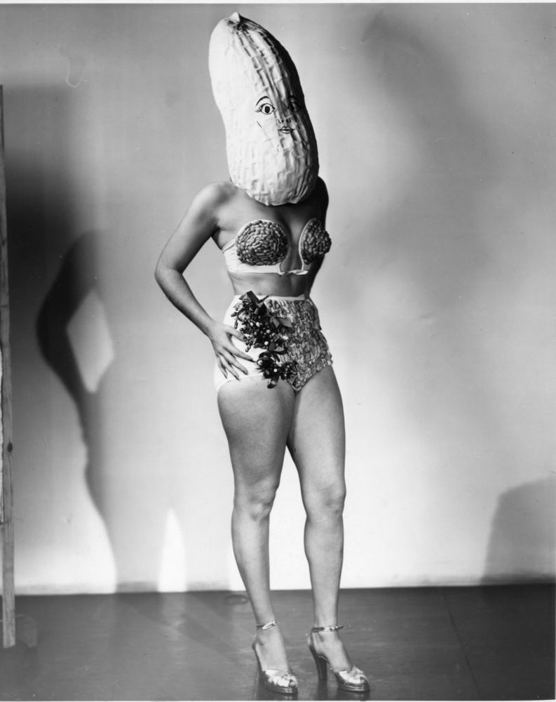 Black and white photo with harsh studio lighting on a woman in a two-piece swim suit that is festooned with sewed-on peanuts, she wears an oversized peanut mask. This is a publicity photo for the National Peanut Festival in Dothan, Alabama, from approximately 1938-1942.