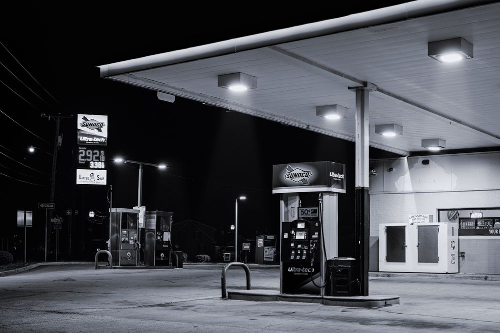 Scene of a gas station at night. The gas pumps are brightly illuminated by overhead lights. To the right is an ice machine and a neon sign showing there is an ATM machine inside the convenience store. To the left are diesel pumps and a tall advertising sign showing the cost of gas. In the background is a donation box for clothing and an air pump for tires. Beyond that, the night has taken over and nothing is visible.