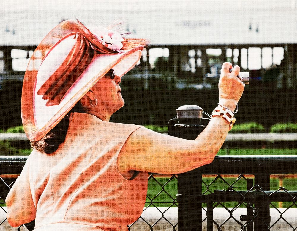 Photo from the 2015 Kentucky Derby. A fashionably dressed woman in a traditional derby bonnet leans over a metal fence rail to snap a photo of the race horses that just passed by on their way to the finish line.