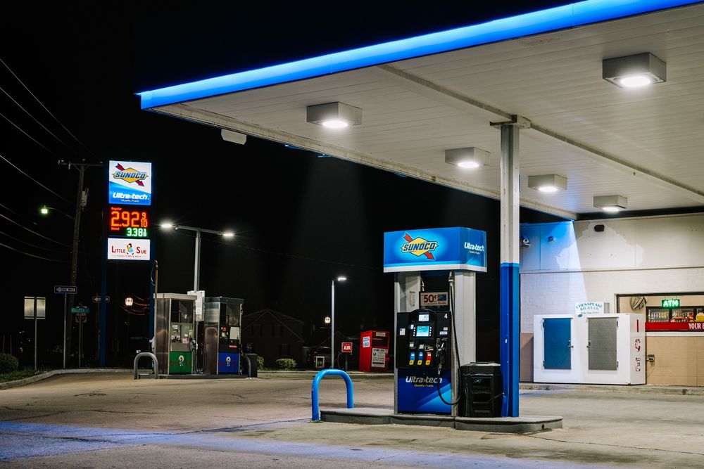 Scene of a gas station at night. The gas pumps are brightly illuminated by overhead lights. To the right is an ice machine and a neon sign showing there is an ATM machine inside the convenience store. To the left are diesel pumps and a tall advertising sign showing the cost of gas. In the background is a donation box for clothing and an air pump for tires. Beyond that, the night has taken over and nothing is visible.