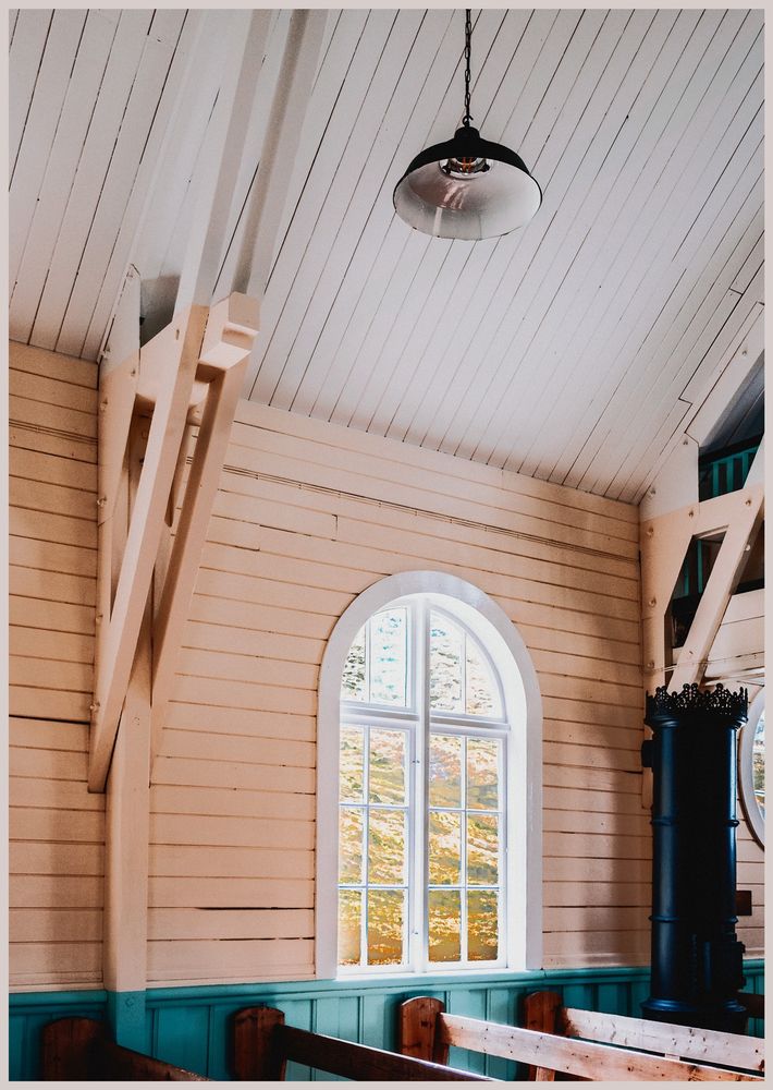 The interior of the Norwegian Anglican Church in Grytviken, South Georgia. The church is a heritage site from when it served the whaling station located on the island. The view looks over the pews to a high arched window. Inside the church is a large fire stove for heating. Outside is a mixture of grass and mud turning to snow as the window reveals the mountain slope beside the church. 