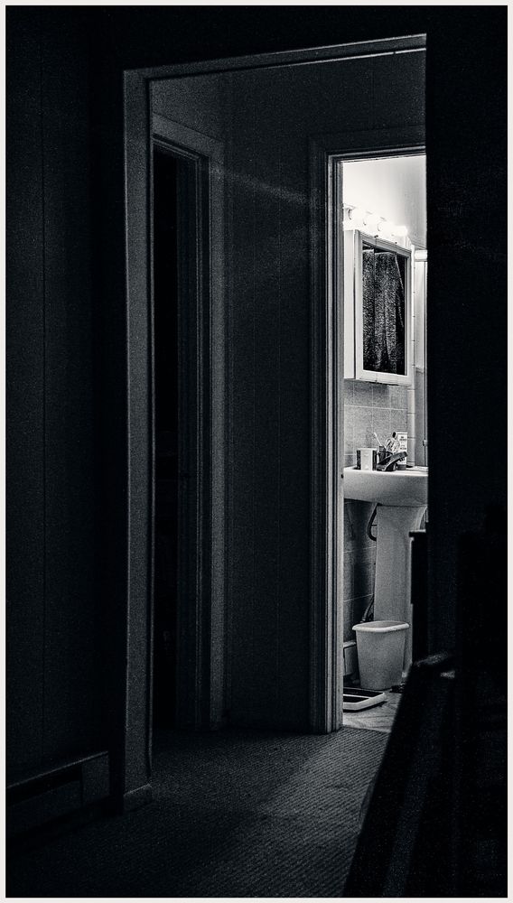 A night scene inside a small house. Bright lights above a pedestal sink brightly illuminate a bathroom. The bulbs project a stream of light into the darkened hallway and a room of the house. Although the illumination is vivid, the gradient of light is quickly absorbed by the moody darkness.
