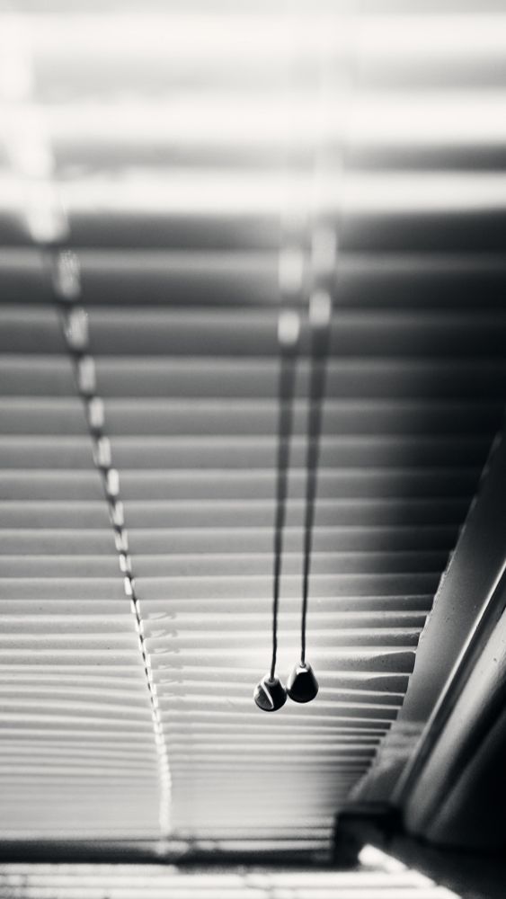Looking down the drawstring of a window blind. The perspective and shallow depth of field show only the two plastic pulleys in focus. Sunlight tries to penetrate the room but the slats prevent its entry.